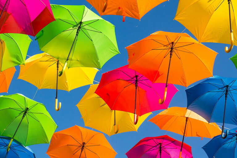A close up of 13 brightly coloured umbrellas (neon green, orange, pick and yellow) hanging on strings with a blue sky in the background.
